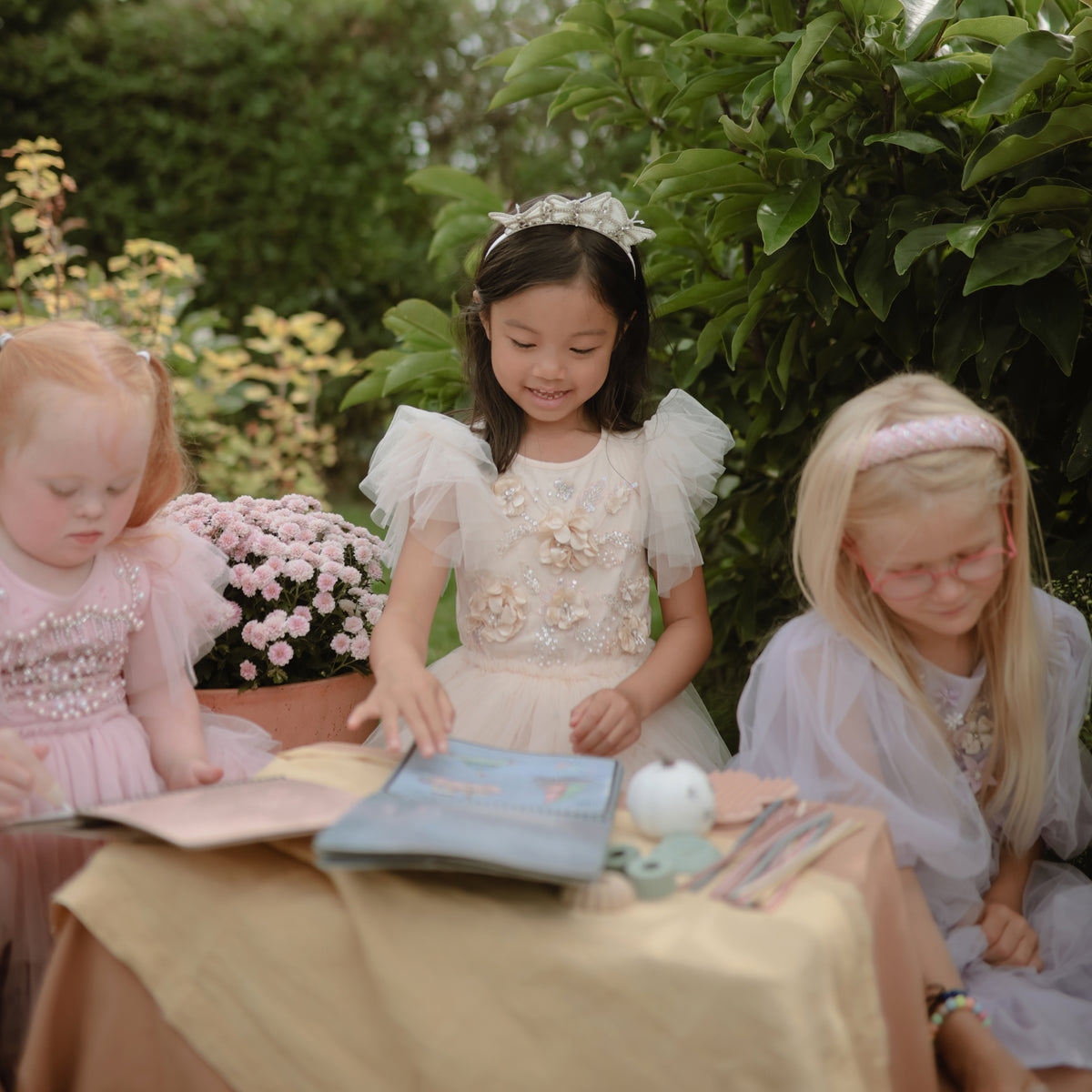 Three girls use mushie’s Reusable Water Color Pad at a small outdoor table in princess dresses.