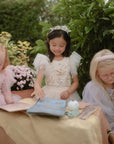 Three girls use mushie’s Reusable Water Color Pad at a small outdoor table in princess dresses.