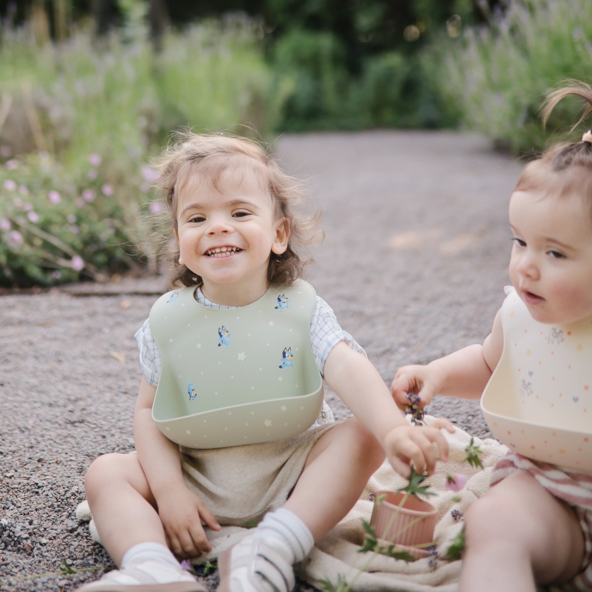 Two toddlers smile on a path with flowers, enjoying the mushie Bluey Gift Set essentials.
