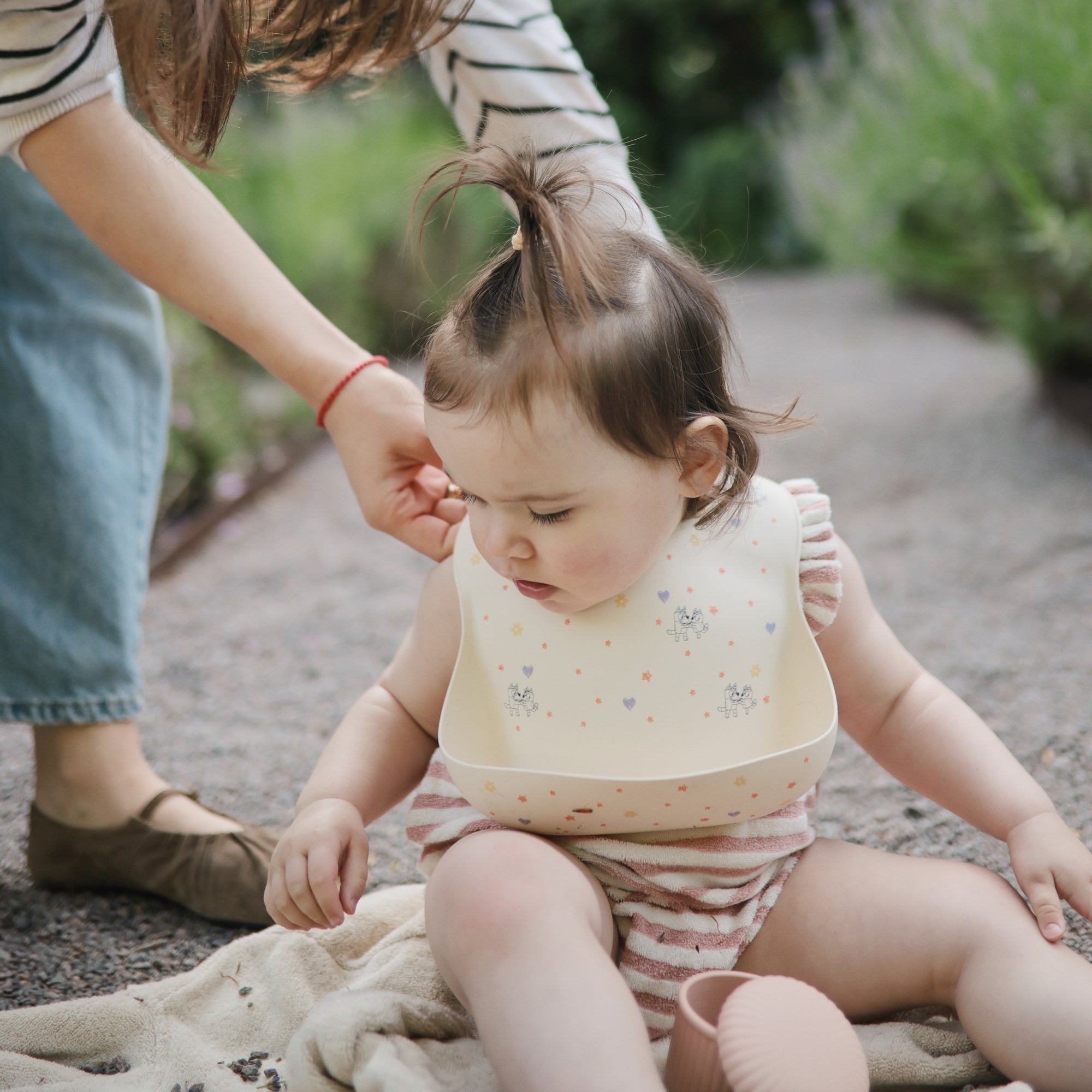 Woman adjusts bib on baby outdoors, using mushie Bluey Gift Set on a blanket.