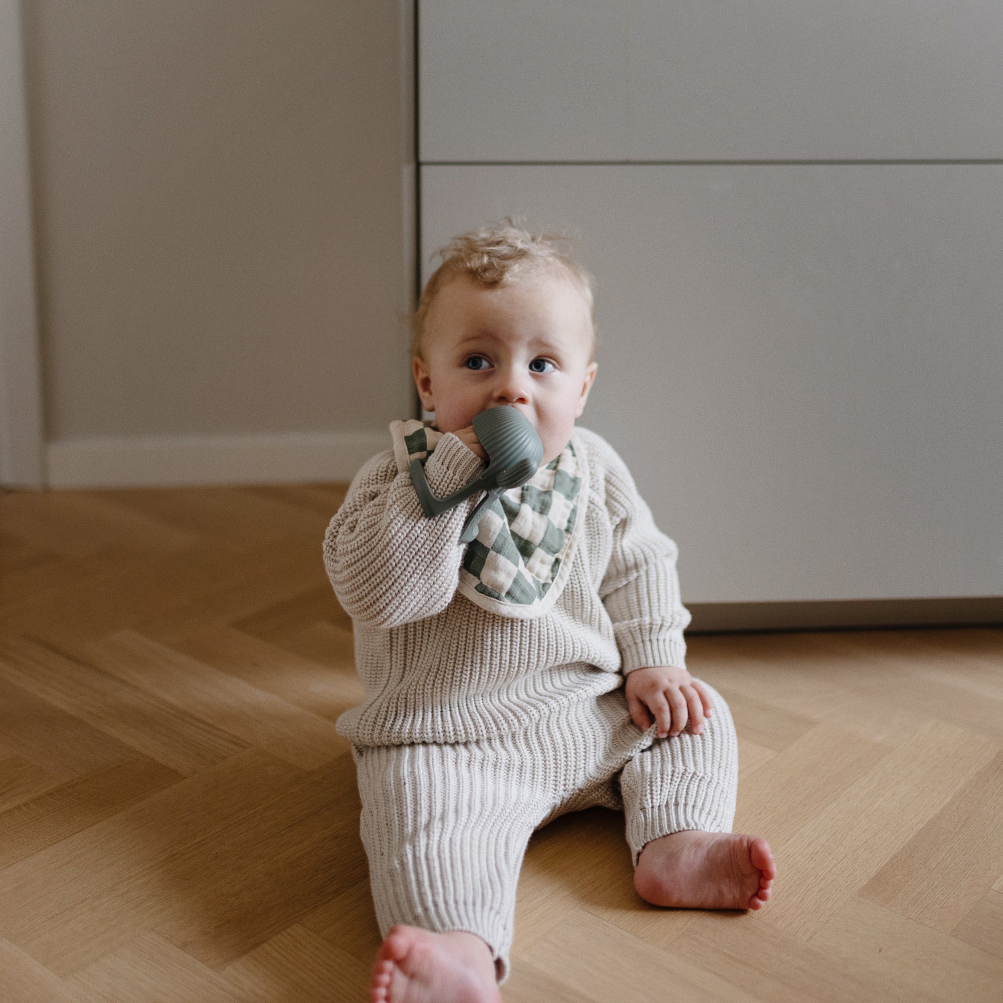 Baby on wooden floor wears a bib and chews a mushie No-Drop Teether in a knit outfit.