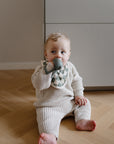 Baby on wooden floor wears a bib and chews a mushie No-Drop Teether in a knit outfit.
