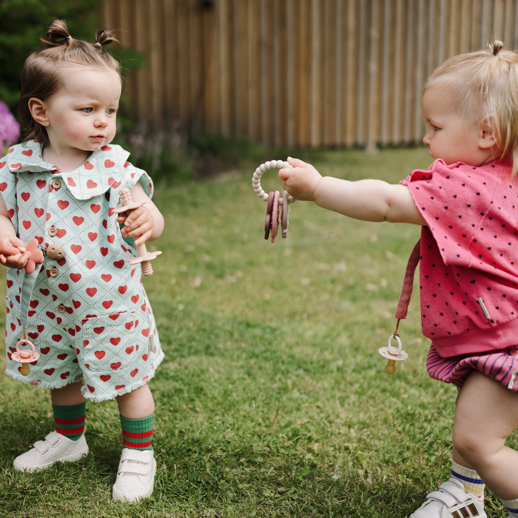 Two toddlers on grass, one gives a mushie Teething Ring to the other.