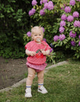 Baby with FRIGG Andersen Fairytale pacifier stands on grass by purple flowers, in pink outfit.