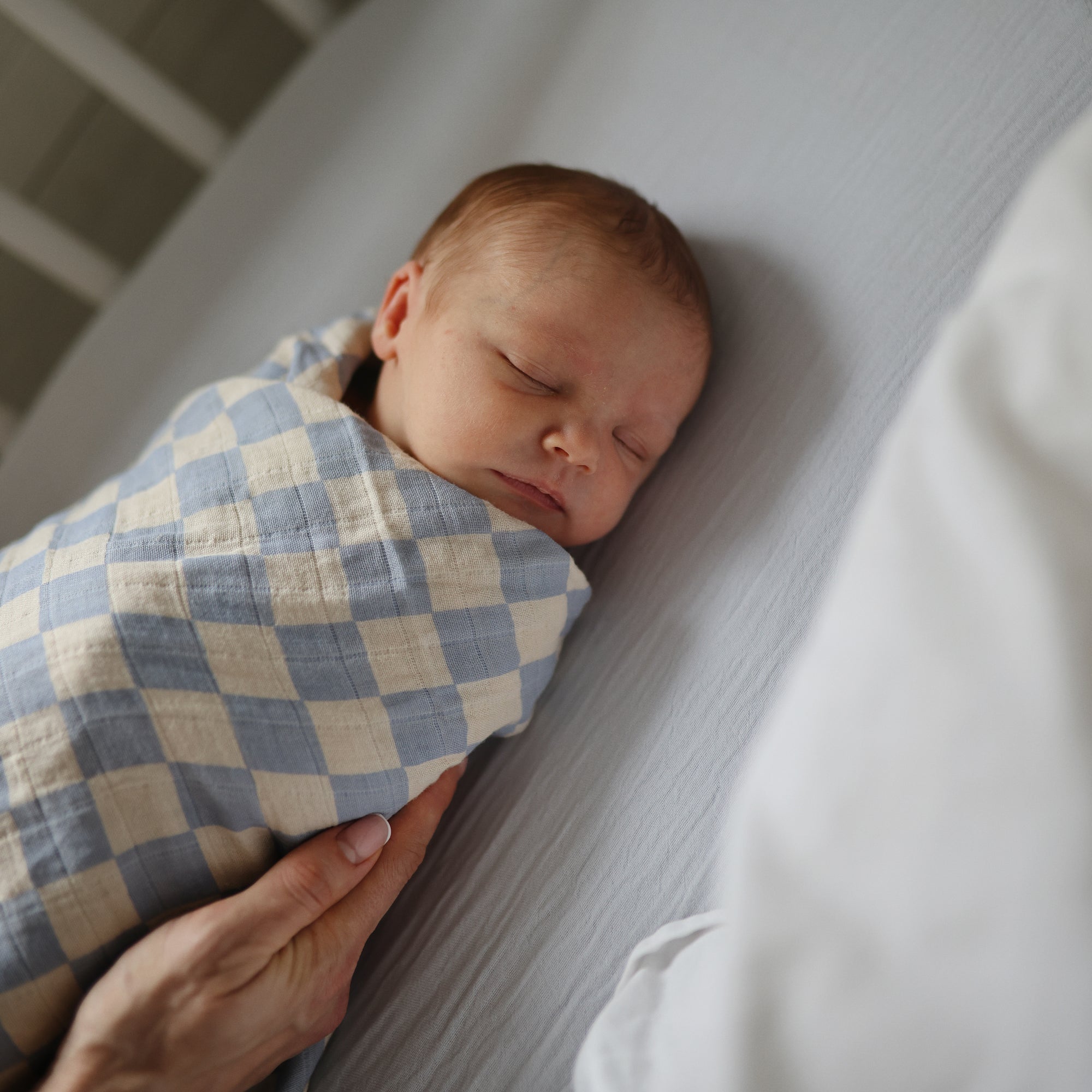 A newborn sleeps peacefully on a light-colored bed with the mushie Extra Soft Bassinet Muslin Crib Sheet, swaddled in a blue and white checkered blanket as an adult hand gently offers comfort.