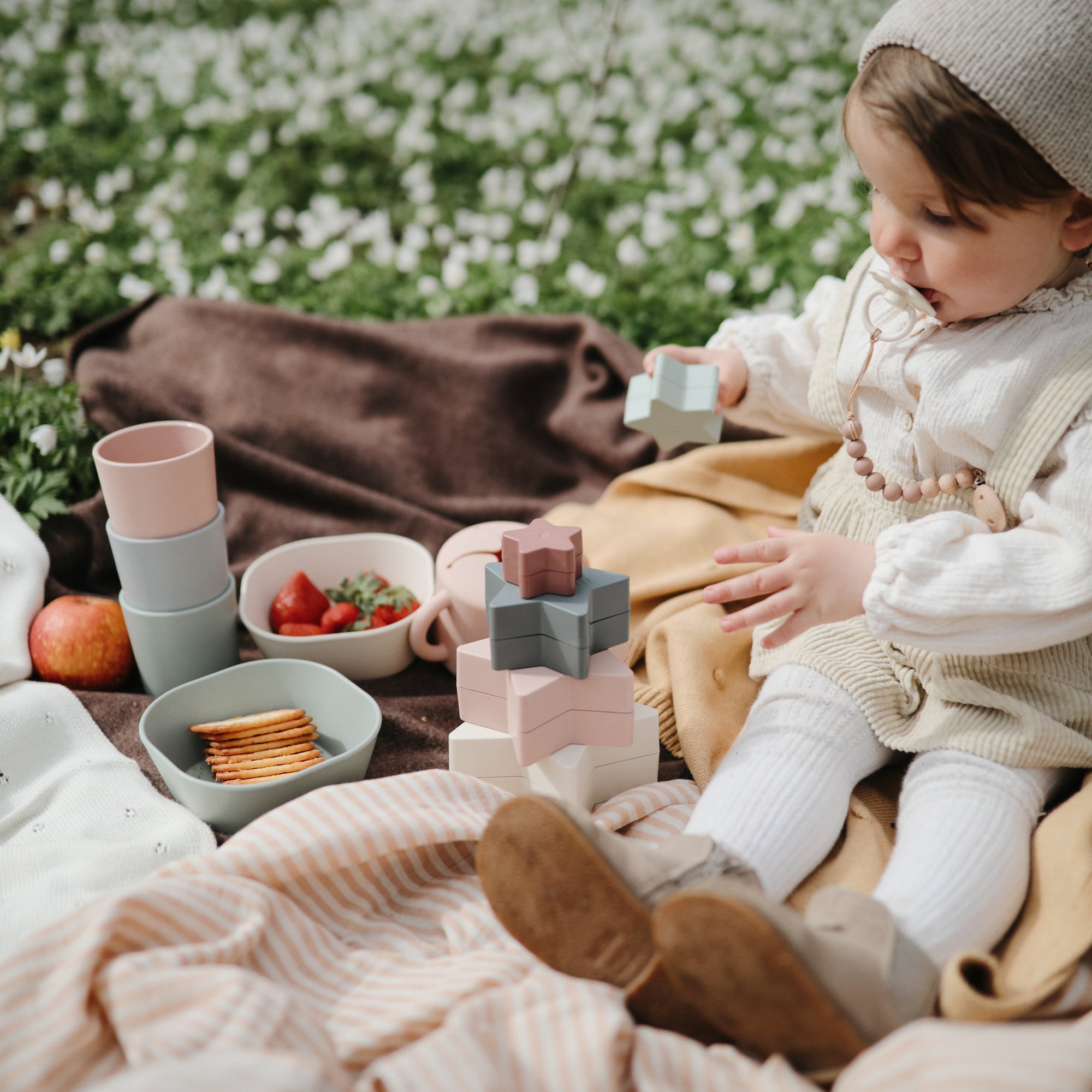 Toddler with stacking toys and a FRIGG Daisy Natural Rubber Pacifier at an outdoor picnic.