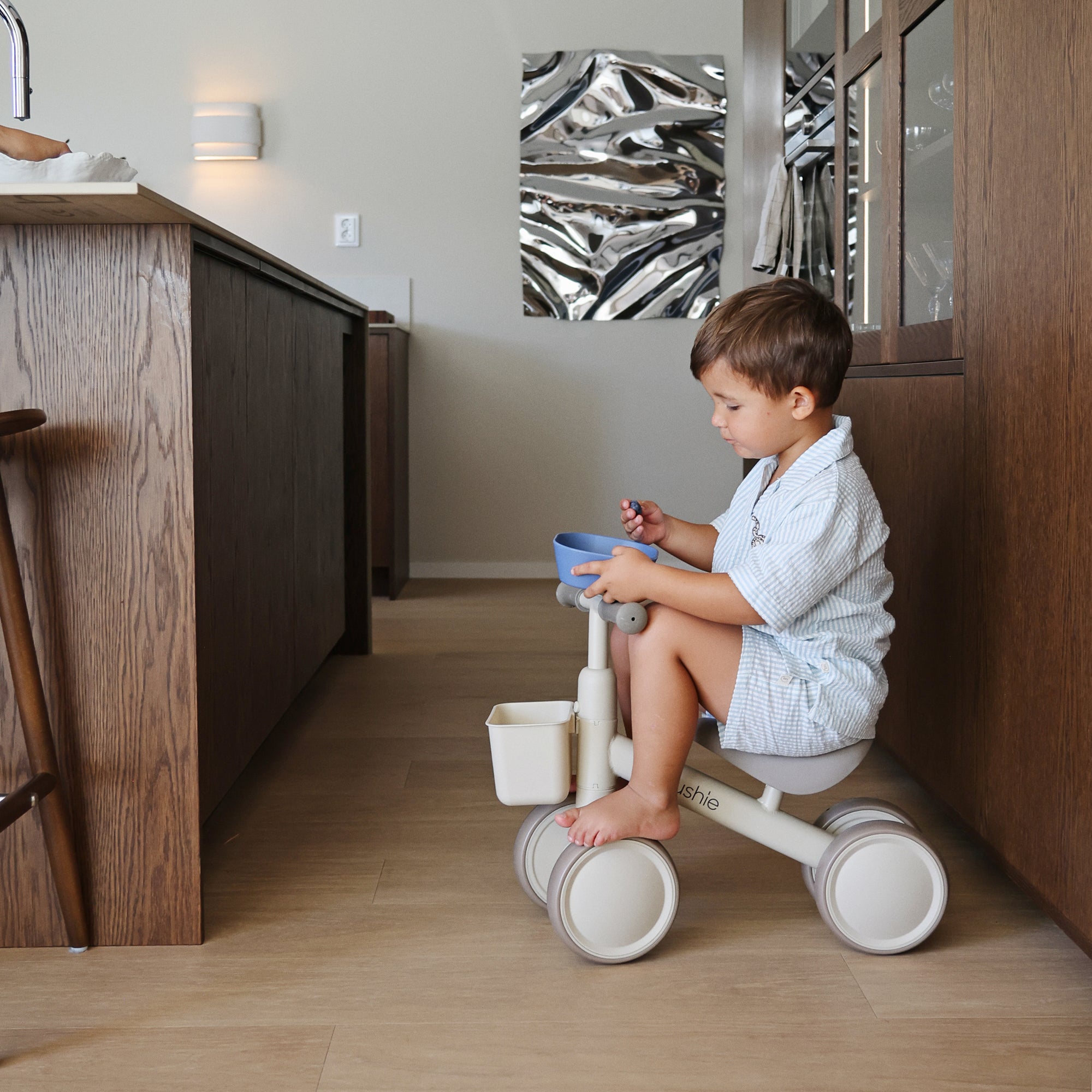Young boy on a tricycle indoors, near a wooden counter with mushie Square Dinnerware Bowls.