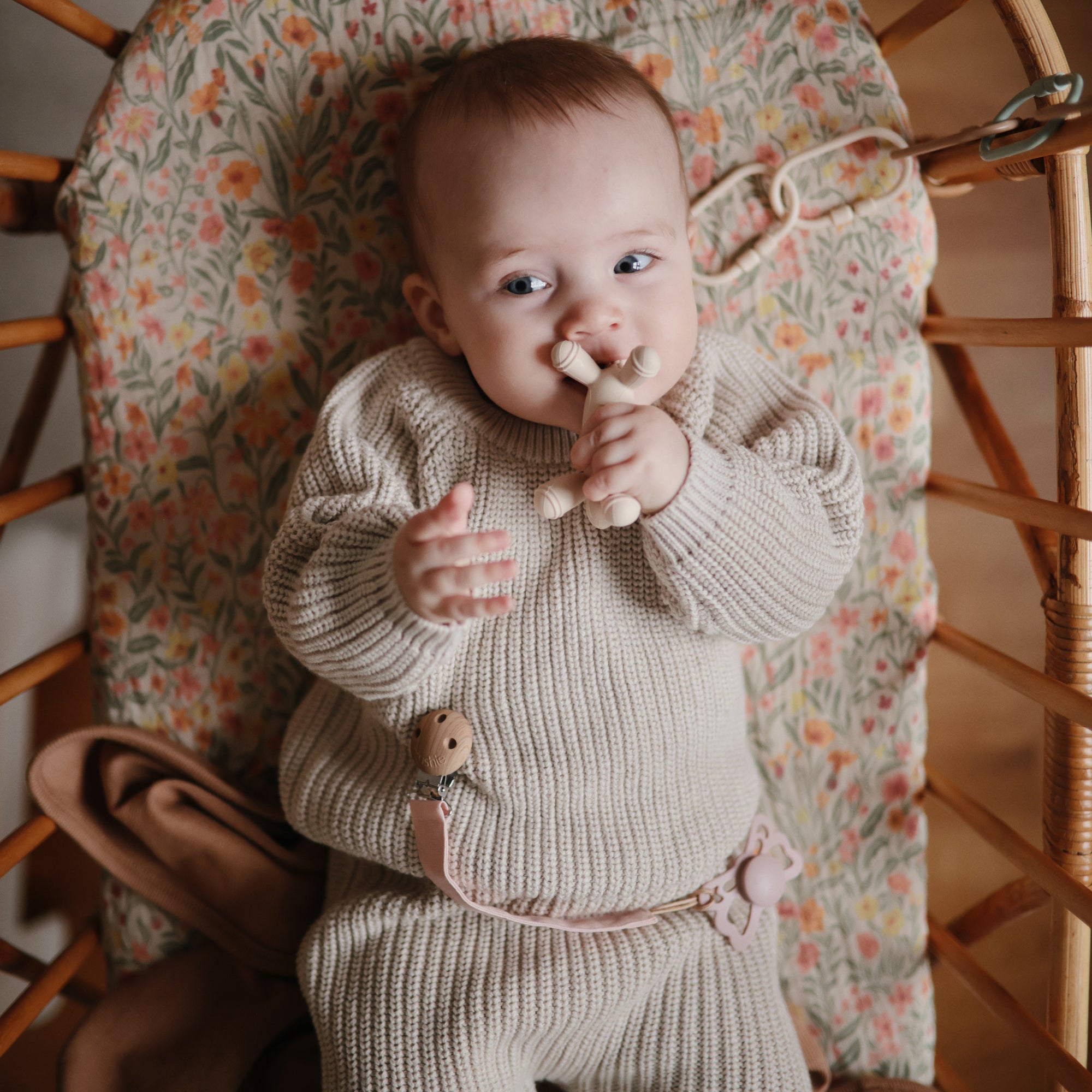 Baby in beige knit outfit with mushie Figurine Teether, lying in floral bassinet.