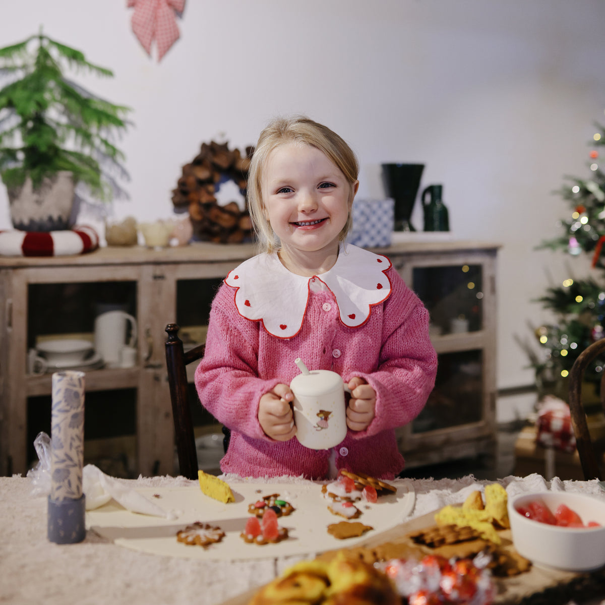 Smiling child holds Mushie Silicone Training Cup + Straw at a table with cookies, holiday decor.