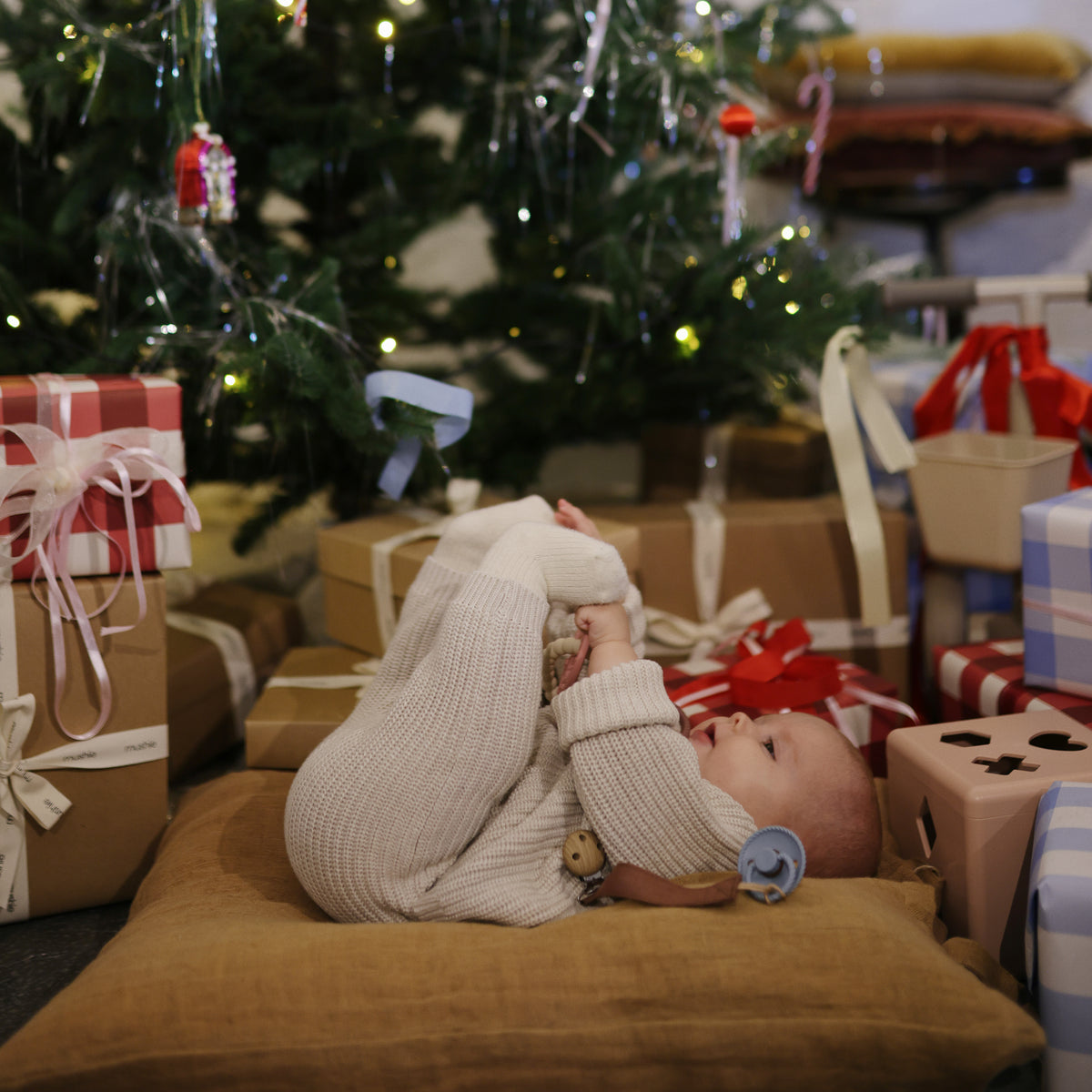 Baby with FRIGG Rope pacifier by Mushie lies on cushion near gifts under a Christmas tree.