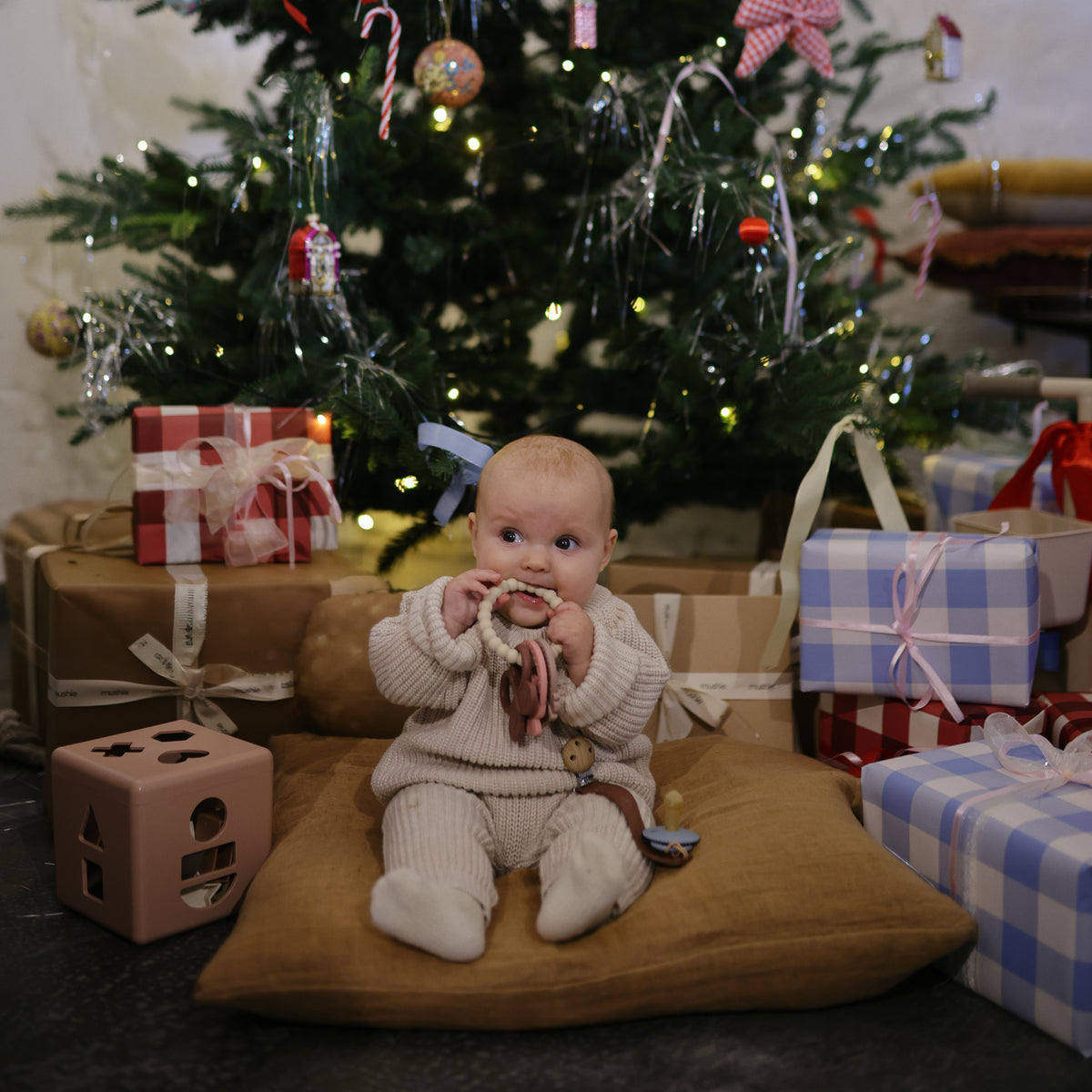 Baby enjoys mushie Teething Ring amid Christmas gifts beneath the festive tree.