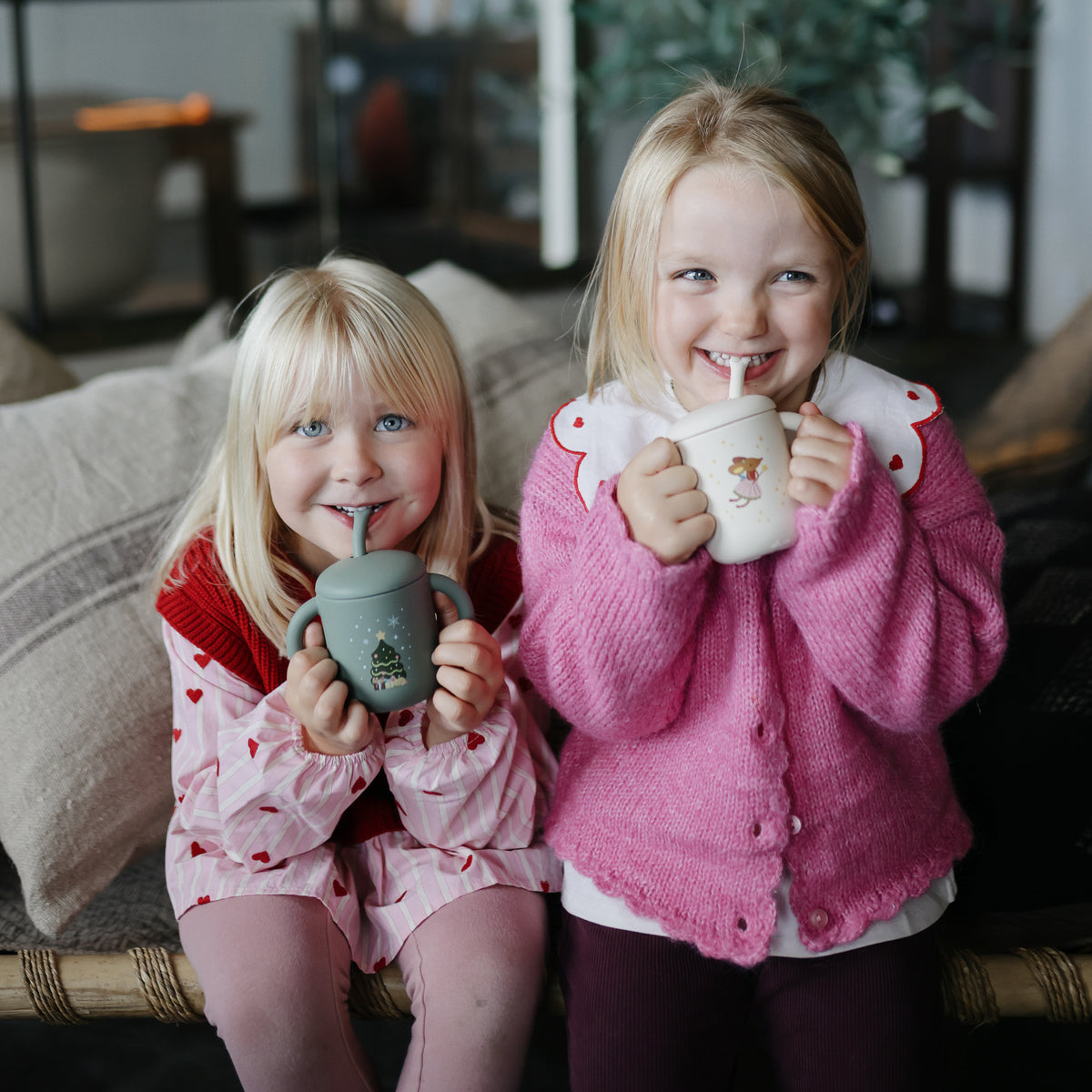 Two smiling girls enjoy sipping from Mushie Silicone Training Cups + Straws.