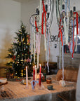 A table with mushie Stacking Cups, candles, and ribbons, a Christmas tree and lights behind.