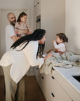 Lifestyle image of a young family of four gathered around a kitchen counter with a young boy sitting on a changing pad  with his mother leaning down and smiling at him. A Cambridge Blue Star Training Toothbrush and Blush Dice Press toy are laid by the baby. 